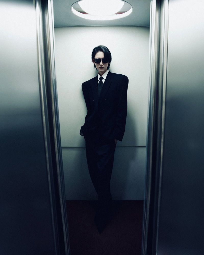 An adult wearing a black suit and sunglasses stands inside of an elevator with mirrored walls and bright overhead lighting.