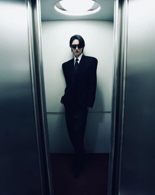 An adult wearing a black suit and sunglasses stands inside of an elevator with mirrored walls and bright overhead lighting.