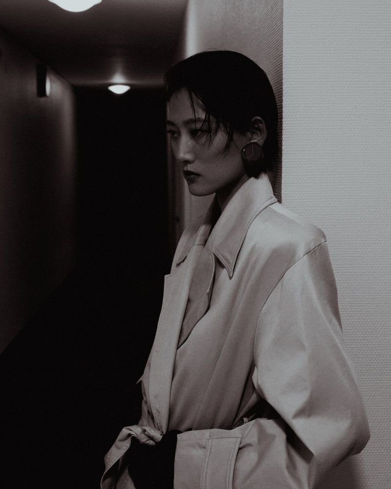 Asian woman in a trench coat stands in a dimly lit hallway, posing for a fashion portrait with a melancholic expression.
