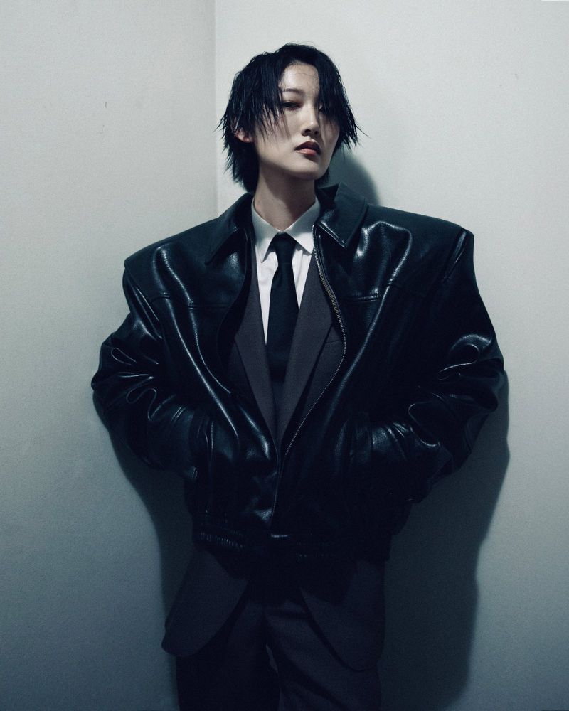 Person in a black leather jacket and grey suit posing against a corner wall, creating a high-fashion, editorial portrait.