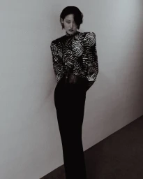 Model with short dark hair in a patterned high-neck top and black trousers leaning against a plain wall.