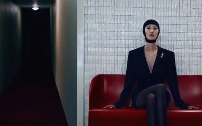 Model in a black hooded balaclava and structured black blazer seated on a red sofa in front of a pale tiled wall and dark hallway.