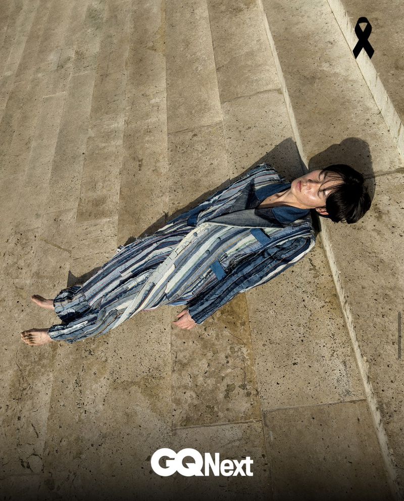 A person with short dark hair lies barefoot on stone steps, wearing a long patchwork denim coat with vertical stripes in various blue tones.