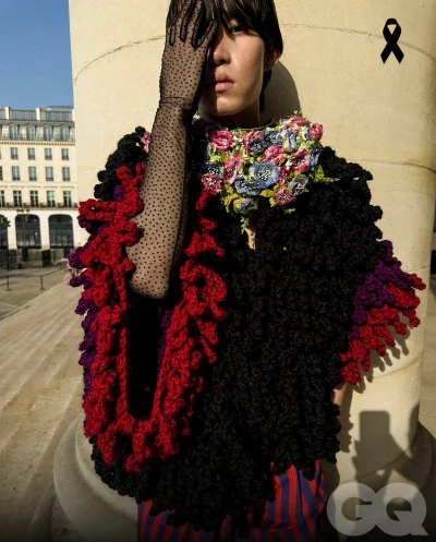 Model Ikko Ohira poses against a stone pillar wearing a black and red textured loop-knit cape and a sheer polka dot glove.