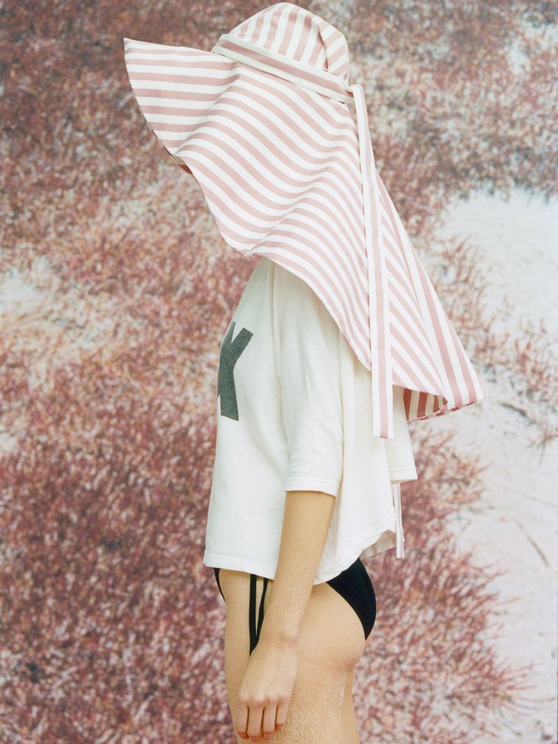 Model in profile wearing an oversized striped sun hat, white t-shirt, and black swim bottoms against a textured backdrop.