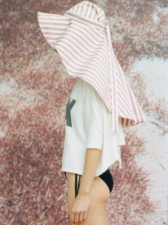 Model wearing an oversized pink and white striped sunhat, white t-shirt, and black bikini bottoms against a textured backdrop.