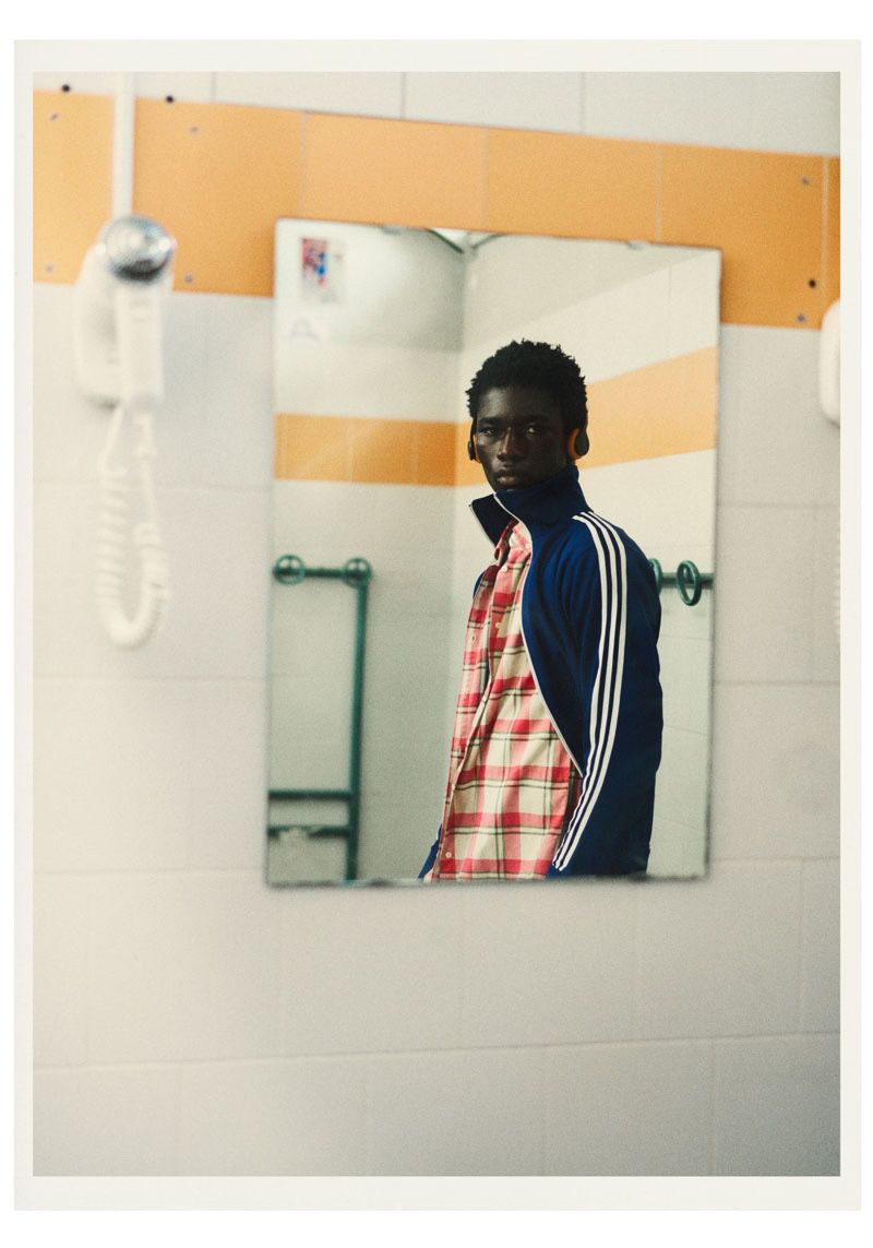 A young Black man wearing a navy blue track jacket over a red plaid shirt, reflected in a rectangular wall-mounted mirror.