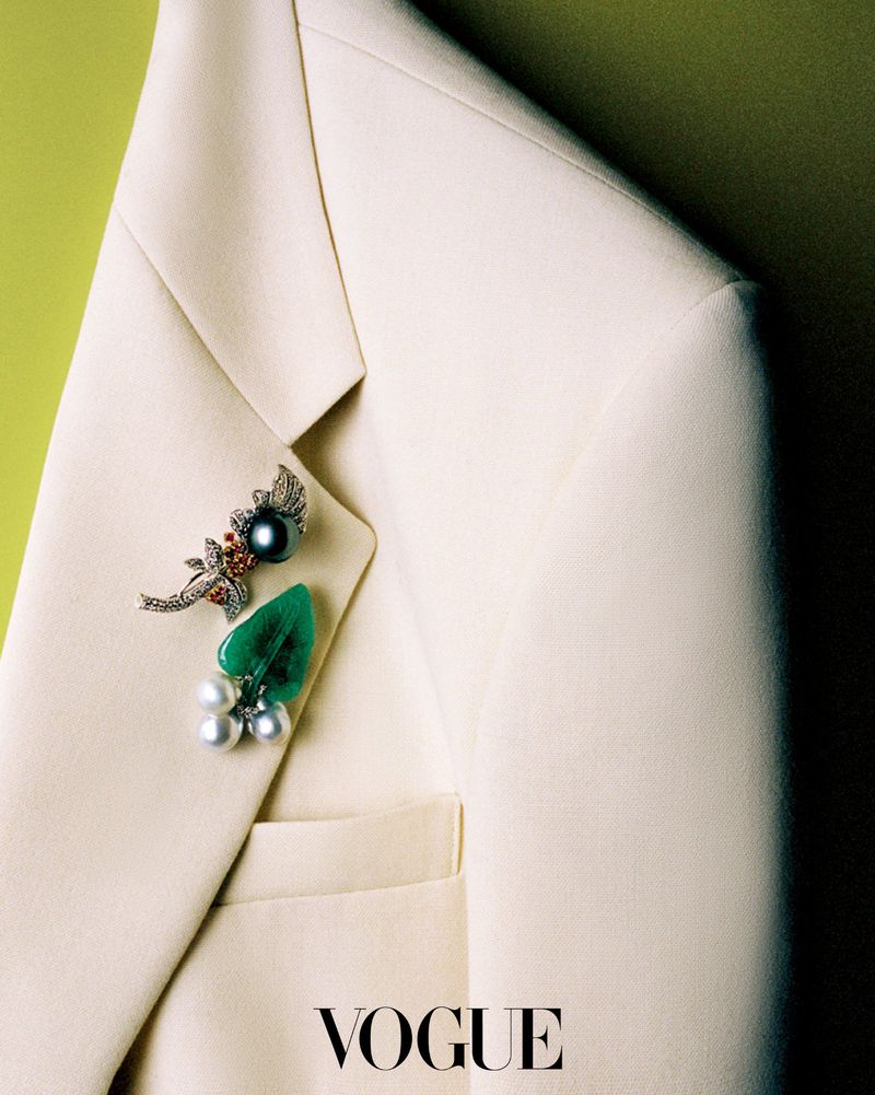 Close-up of ornate pearl and gemstone brooches pinned to the lapel of an off-white tailored blazer.