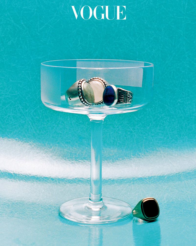 Clear coupe cocktail glass holding silver and gemstone rings with a green signet ring beside it on a bright cyan surface.