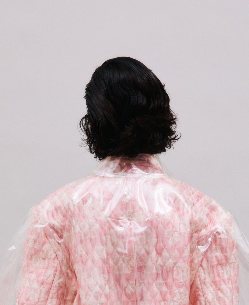 Rear view of a person with short wavy dark hair wearing a quilted pink jacket beneath a clear vinyl overlay against a grey backdrop.