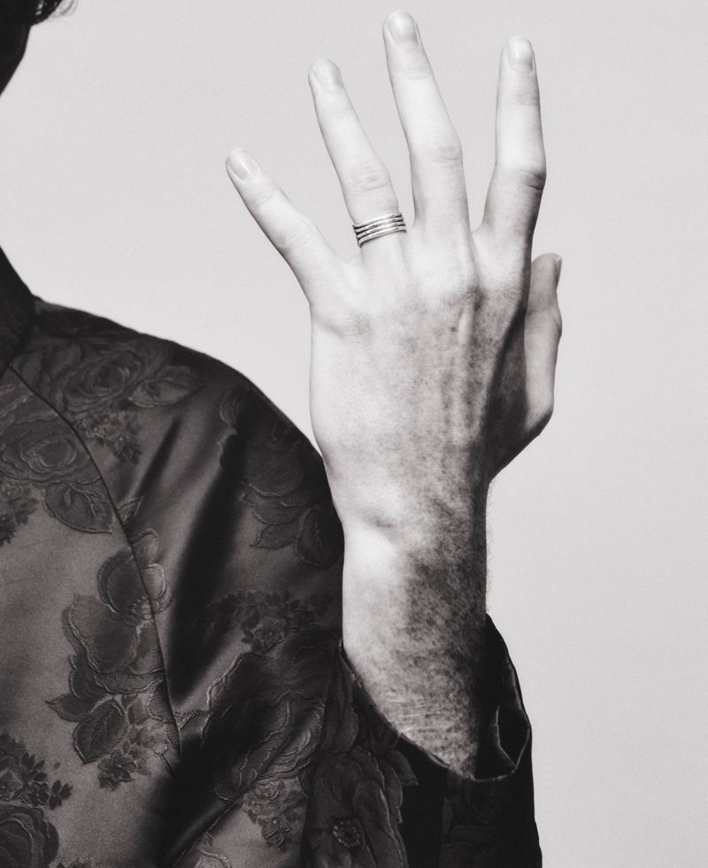 Close-up black and white photograph of a raised hand wearing a silver stacked ring and a floral jacquard jacket against a pale backdrop.