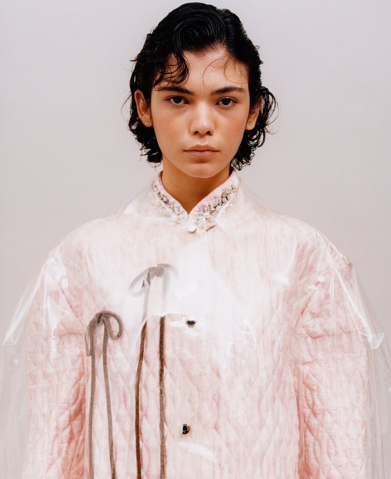 Model wearing a pale pink quilted jacket with an embellished collar and clear overlay against a neutral backdrop.