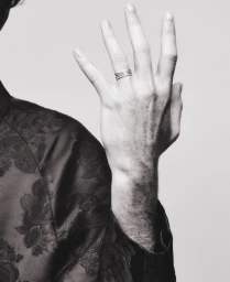 Close-up black and white photograph of a raised hand wearing a silver stacked ring and a floral jacquard jacket against a pale backdrop.