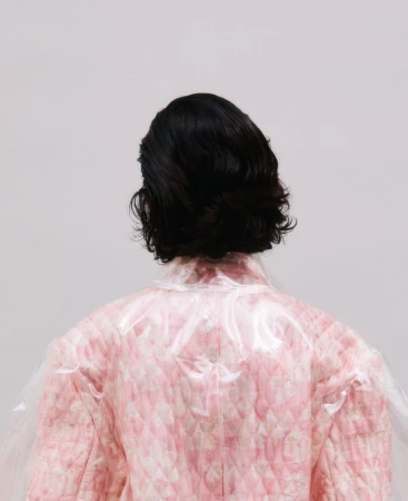 Back view of a person with dark wavy hair wearing a pink quilted jacket under a transparent vinyl coat against a light grey studio backdrop.