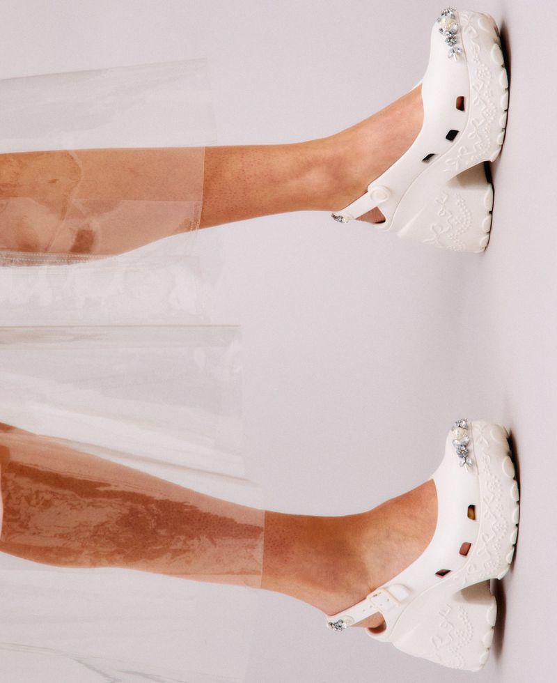 Close-up of legs wearing white crystal-embellished platform clogs and clear transparent socks against a pale studio backdrop.