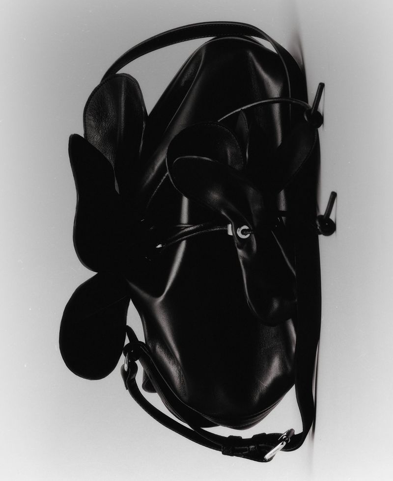 Black sculptural leather handbag with petal-shaped bow detailing against a neutral studio backdrop.