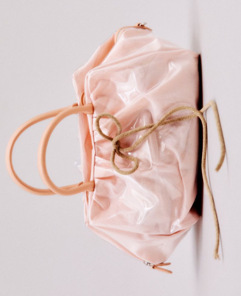 Pale pink glossy ruched handbag with structured top handles and a tan cord bow accent set against a white studio background.