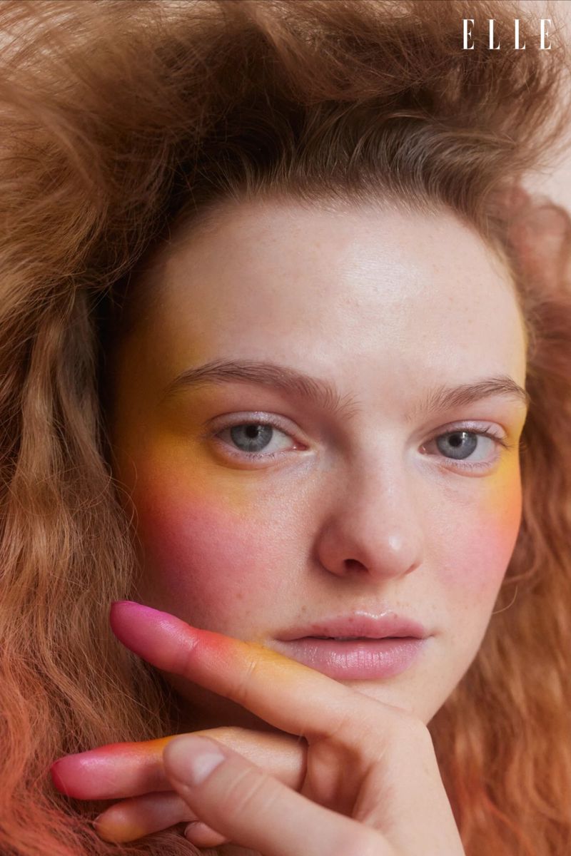Close-up portrait of a fair-skinned adult woman with wavy red hair and artistic makeup in hues of pink, orange, and yellow.