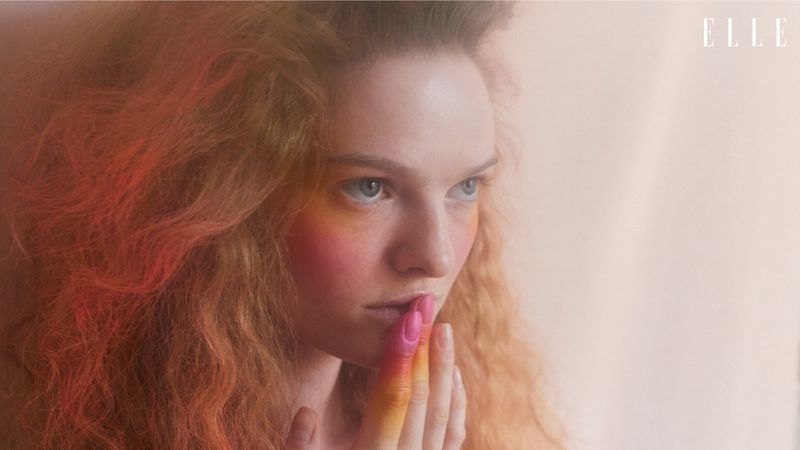 A woman in an orange dress poses against a tan backdrop in a studio, bathed in soft, directional lighting and framed in a medium shot.
