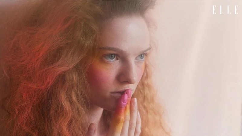 A woman in an orange dress poses against a tan backdrop in a studio, bathed in soft, directional lighting and framed in a medium shot.