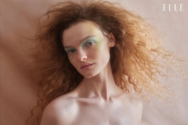 Portrait of a woman with red curly hair and green eyeshadow, featuring a soft focus and natural lighting, against a peach background.
