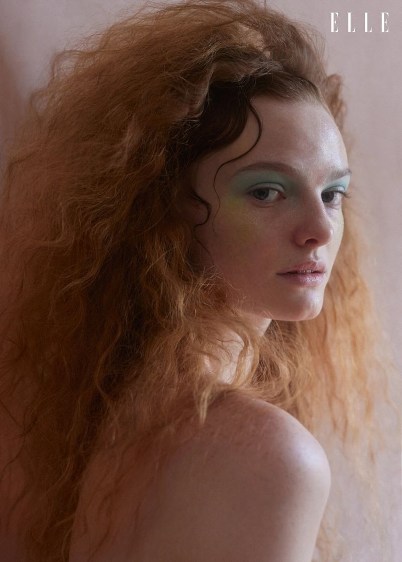 Close-up studio shot of a young adult woman with red curly hair and blue eye shadow, fair skin, partially bare shoulders, and the 'ELLE' logo visible.