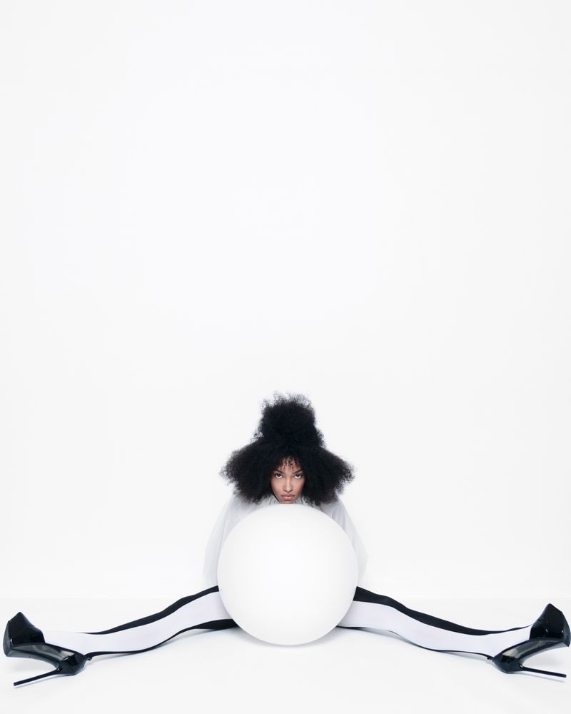 Woman in black afro wig strikes a pose in white studio with a white ball, wearing black and white striped tights, and black high heels.