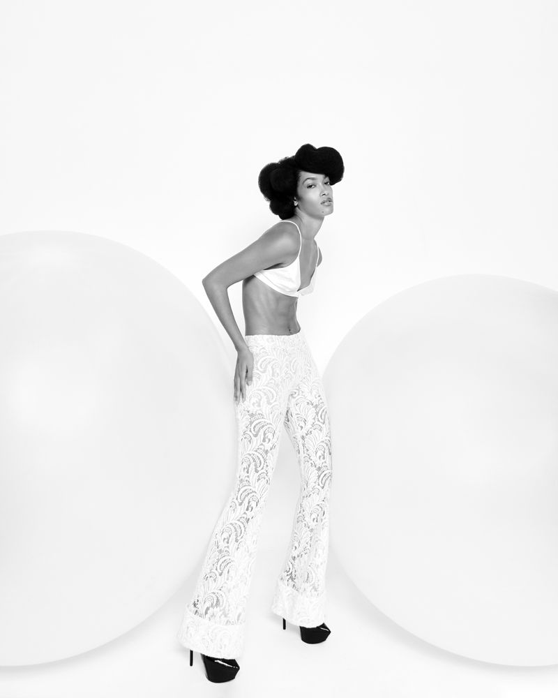 A slender adult female poses in a white bra top and lace pants in a studio with large white orbs, set against a clean white backdrop.