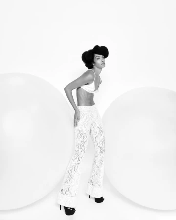A slender adult female poses in a white bra top and lace pants in a studio with large white orbs, set against a clean white backdrop.