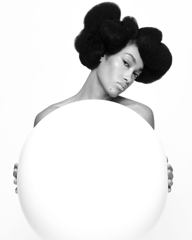 Portrait of a Black woman with a large afro hairstyle, holding a large white sphere against a white backdrop in a studio setting.