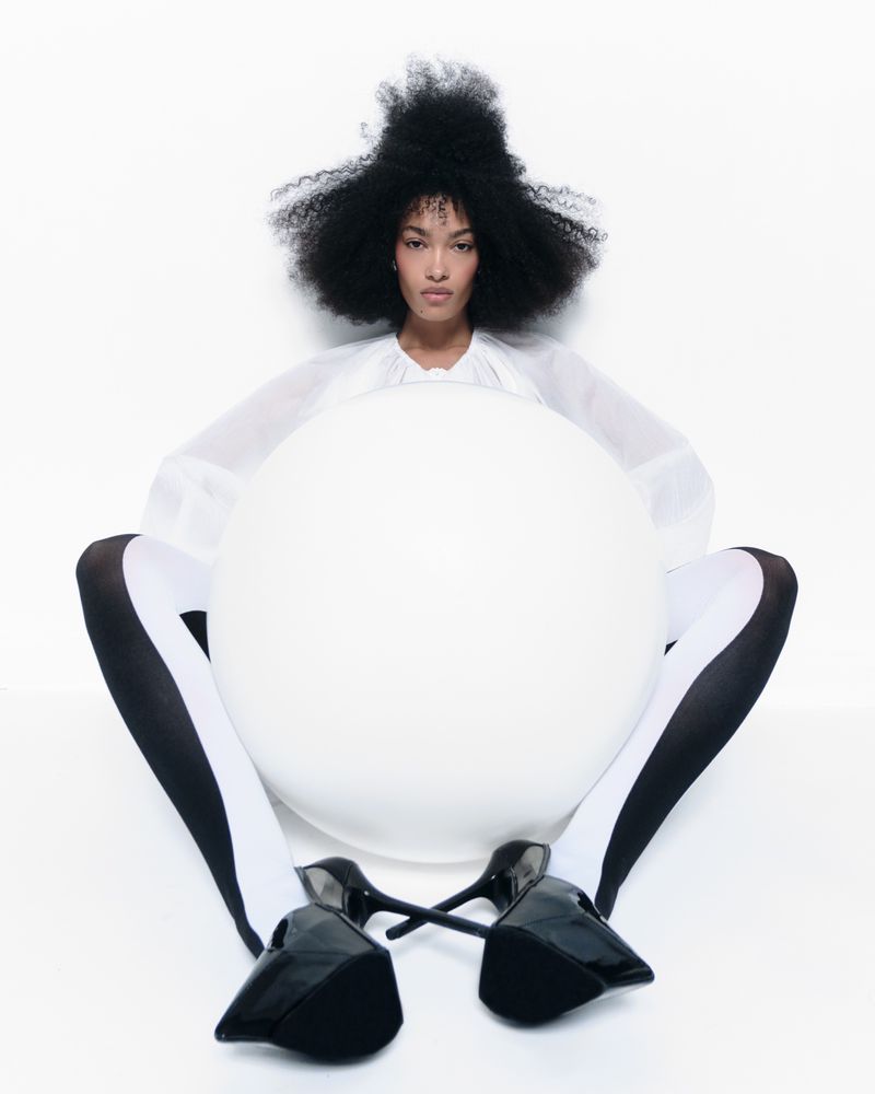 Fashion image featuring an adult female with afro hairstyle, wearing a white top, striped tights, and black high heels in a white studio.