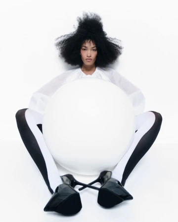 Fashion image featuring an adult female with afro hairstyle, wearing a white top, striped tights, and black high heels in a white studio.