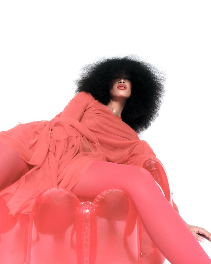 A black woman with an afro hairstyle poses in a red dress and tights while sitting on an inflatable red chair on a white background.