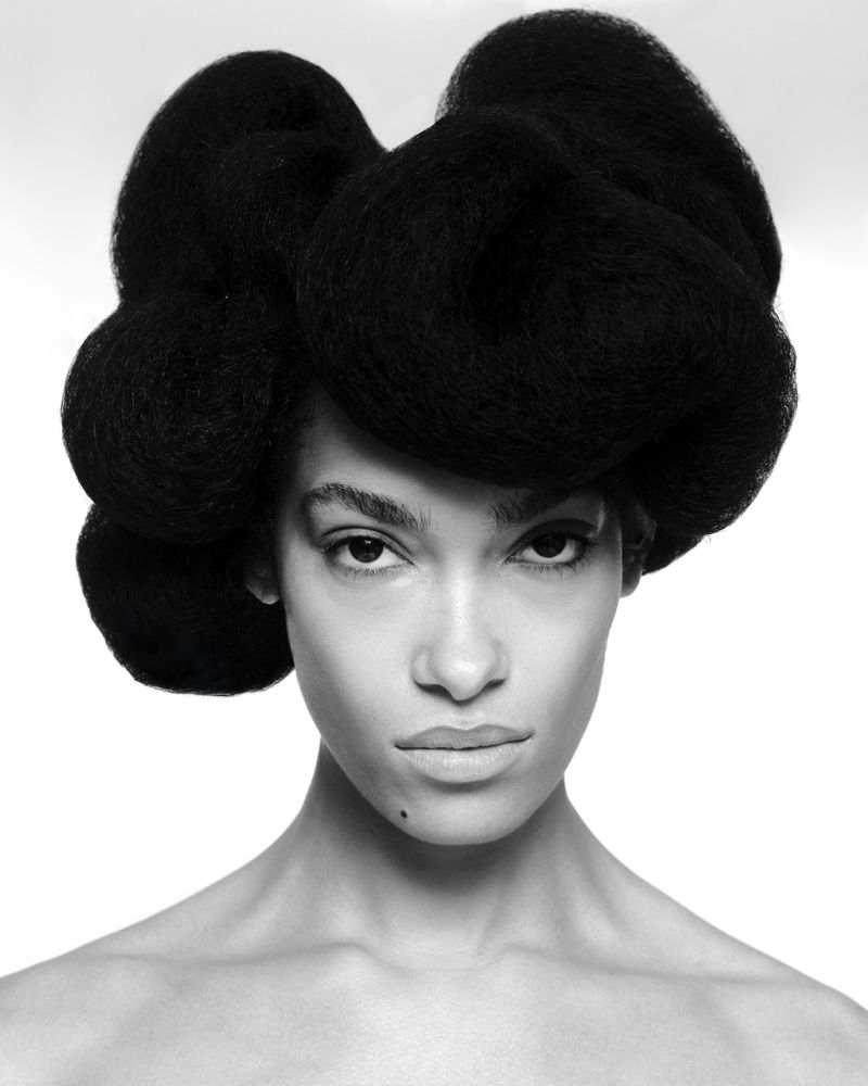 Close-up portrait of a Black woman with dark hair styled in a large, sculptural shape against a bright white background. 