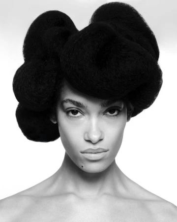 Close-up portrait of a Black woman with dark hair styled in a large, sculptural shape against a bright white background. 