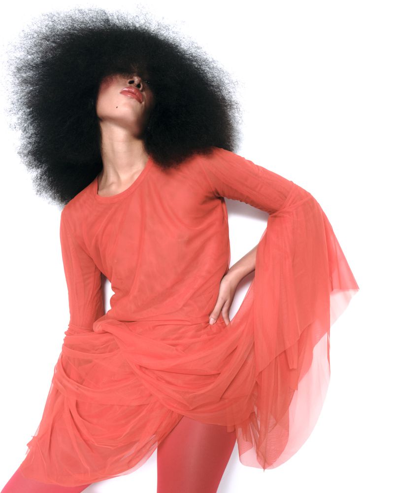 Woman with large afro hairstyle poses in a coral dress and matching tights against a white background in a fashion photography studio.
