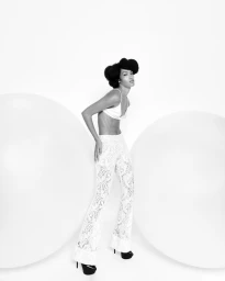 Model standing between two large spherical props wearing patterned white trousers and a white top against a seamless background