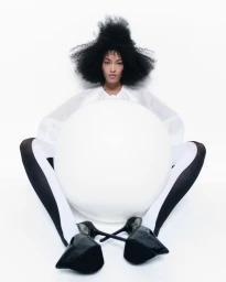 Model with voluminous dark hair holding a large white sphere while wearing a white top and black heels against a white background