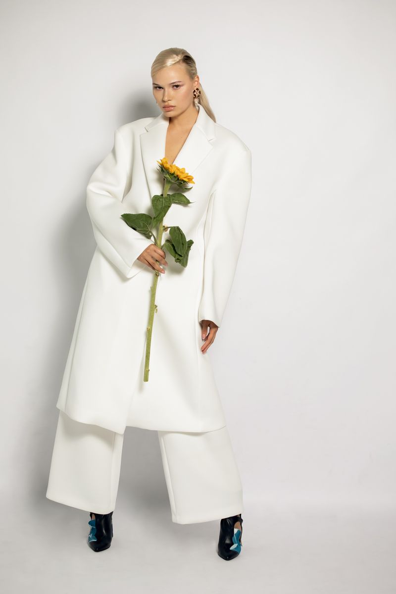 A blonde woman in a white coat holds a sunflower in a studio setting, facing the camera with a neutral expression, showcasing fashion and beauty.