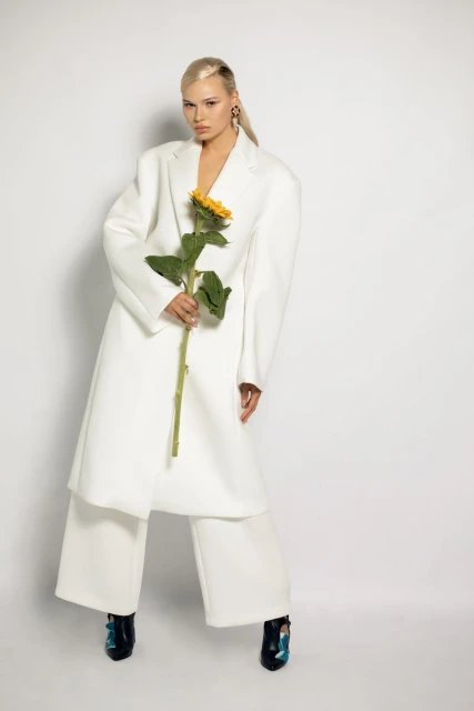 A blonde woman in a white coat holds a sunflower in a studio setting, facing the camera with a neutral expression, showcasing fashion and beauty.