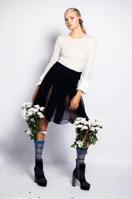 Fashionable woman in stylish clothes and platform boots with flower-filled socks in a studio setting against a white background.