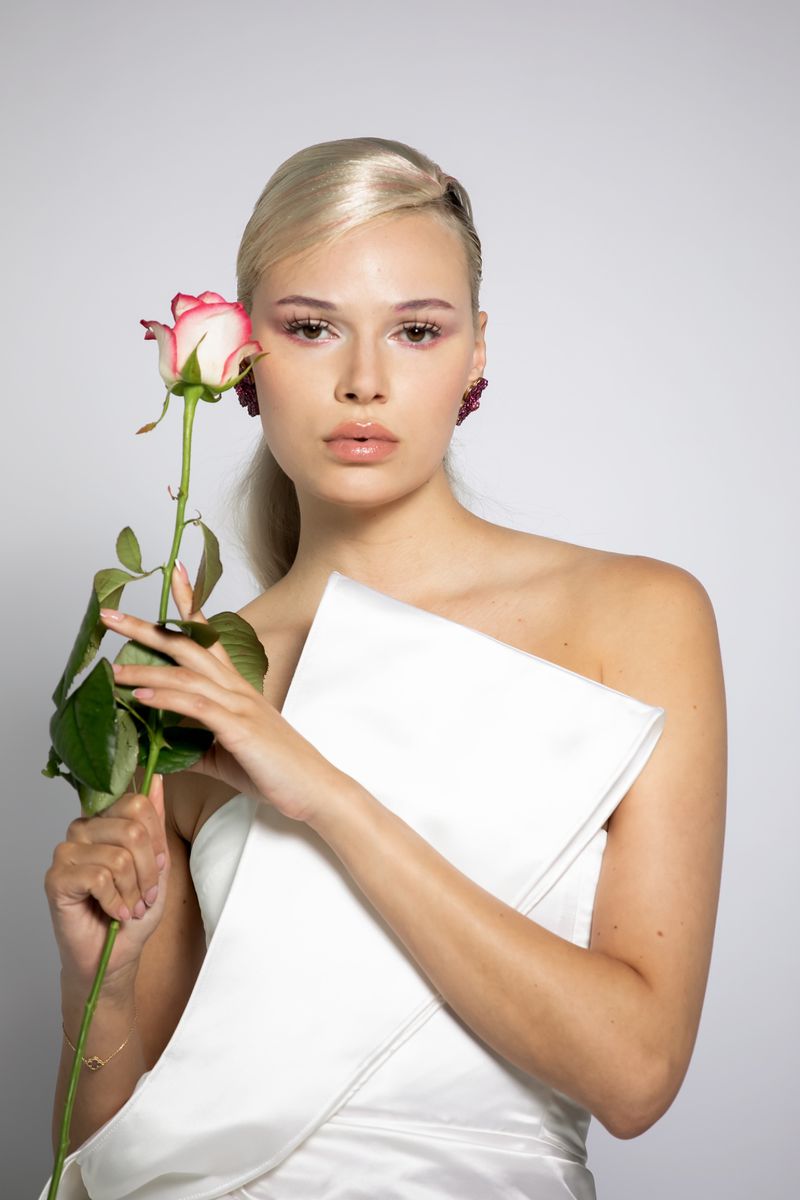 Close-up of an adult female posing with a pink rose in a photo studio setting, wearing elegant white clothing and statement earrings.