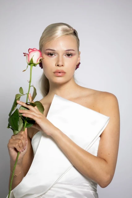 Close-up of an adult female posing with a pink rose in a photo studio setting, wearing elegant white clothing and statement earrings.