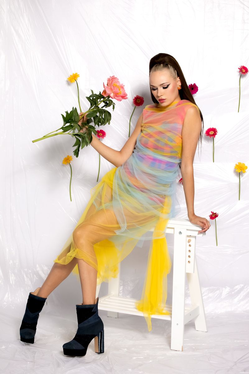 A woman in a colorful dress poses on a white stool with a bouquet of pink flowers in a white photo studio with gerbera daisies.
