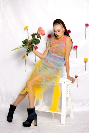 A woman in a colorful dress poses on a white stool with a bouquet of pink flowers in a white photo studio with gerbera daisies.