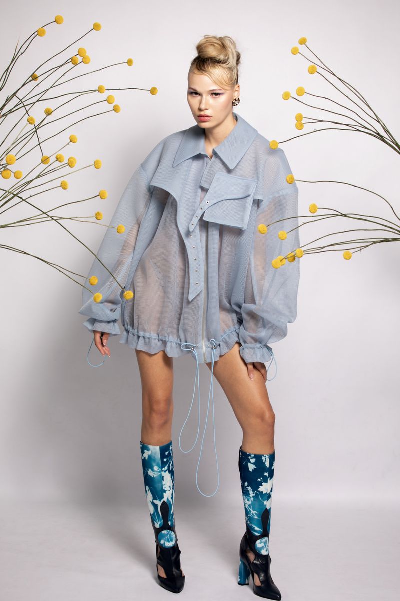 Fashion shot of a female model in an oversized, translucent blue jacket with floral decor, and high-heeled boots on a white background.
