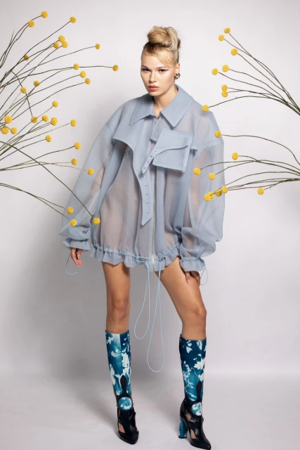 Fashion shot of a female model in an oversized, translucent blue jacket with floral decor, and high-heeled boots on a white background.