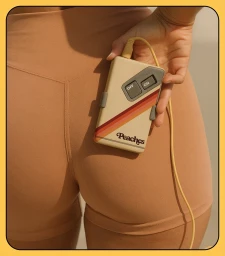 Close-up of a hand holding a vintage-style Peaches branded device against peach-colored athletic shorts.