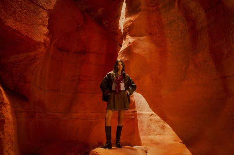 Mathilde Henning standing in a red sandstone slot canyon wearing a dark jacket, mini skirt, and knee-high black boots.