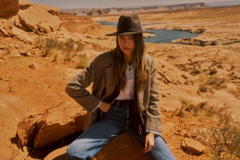 Mathilde Henning seated on a sandstone rock wearing a plaid blazer, fedora hat, and blue jeans in a desert landscape.